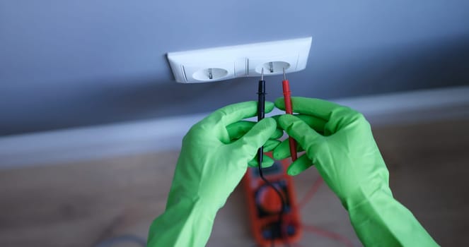 Gloved hands are holding a digital multimeter near the socket, close-up. Test device. AC and DC current measurement