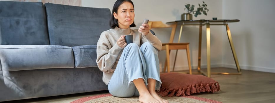 Portrait of young woman with remote, watching tv, switching chanels on television, sitting on floor near sofa and relaxing.