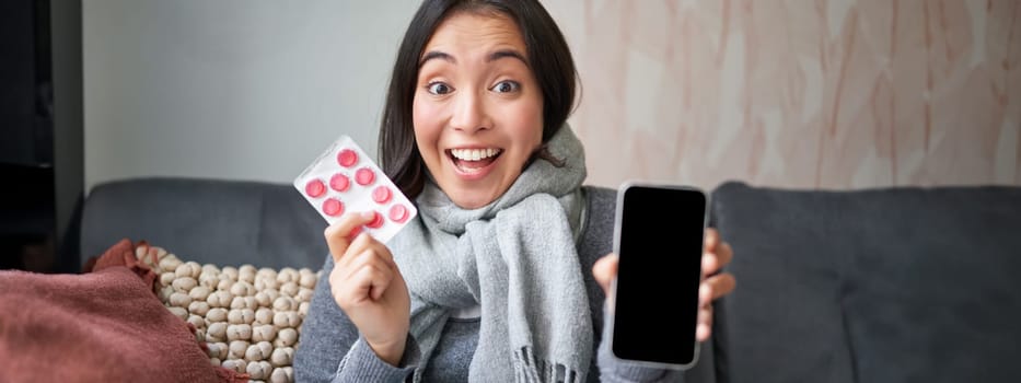 Online doctors and healthcare. Smiling korean woman shows mobile phone screen and medication, catching cold, staying at home and recommending smartphone GP.