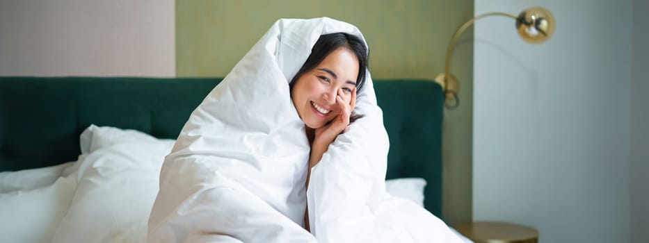 Beautiful asian woman sitting on bed, covered in blankets and duvet, laughing and smiling, enjoying weekend in bedroom.