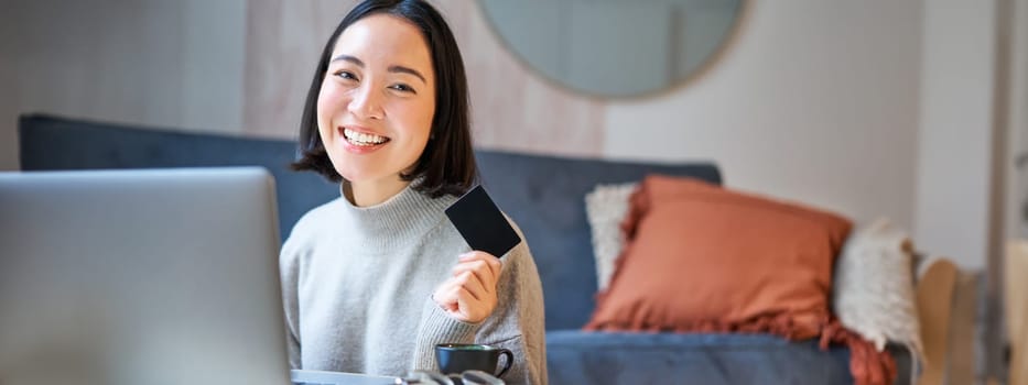 Happy smiling girl with credit card, paying her bills online on computer, doing shopping on her laptop, sitting at home.