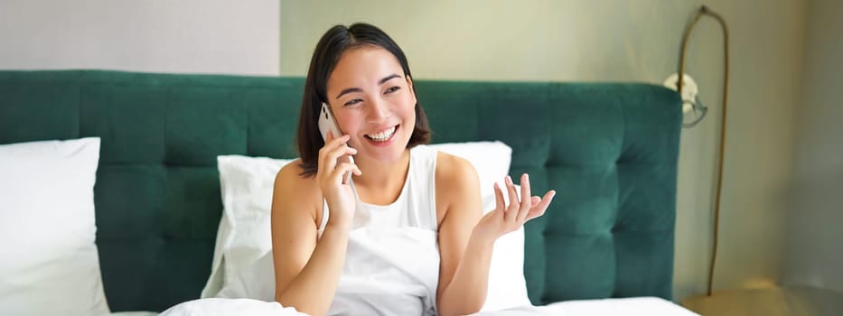 Woman talks on mobile phone in bed. Smiling girl having telephone conversation while relaxing in bedroom.