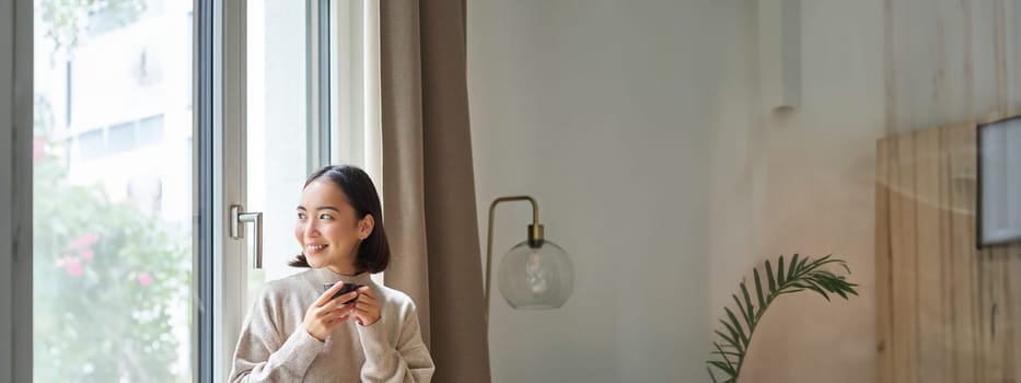 Portrait of young asian woman sitting near window and looking outside, drinking hot coffee from espresso cup and enjoying her cozy day off at home.