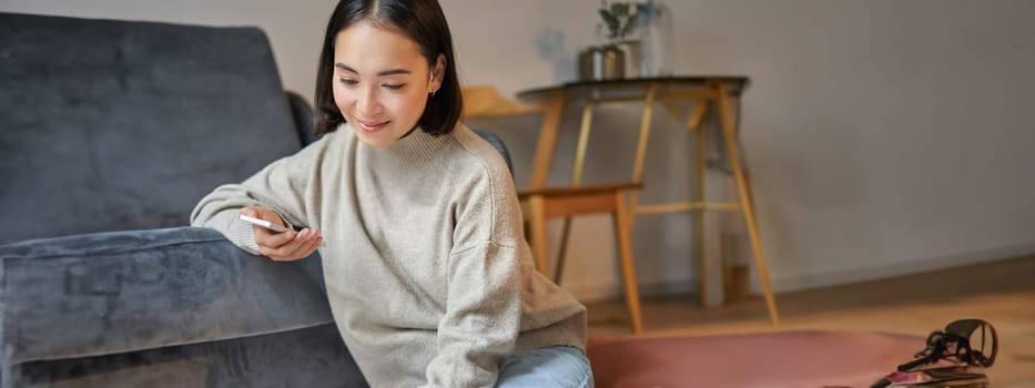 Vertical shot of young woman in cozy home working on laptop, using smartphone and drinking coffee, sitting on floor near sofa.