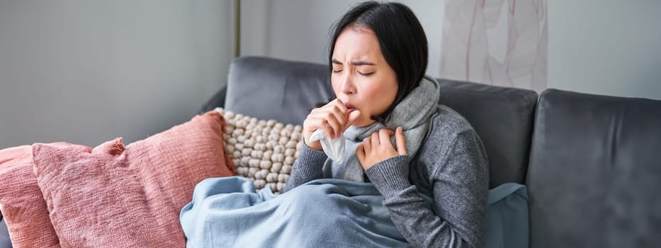 Asian woman coughing, feeling sick, sitting under blanket with scard in cold apartment, saving on heating this winter, catching flu.