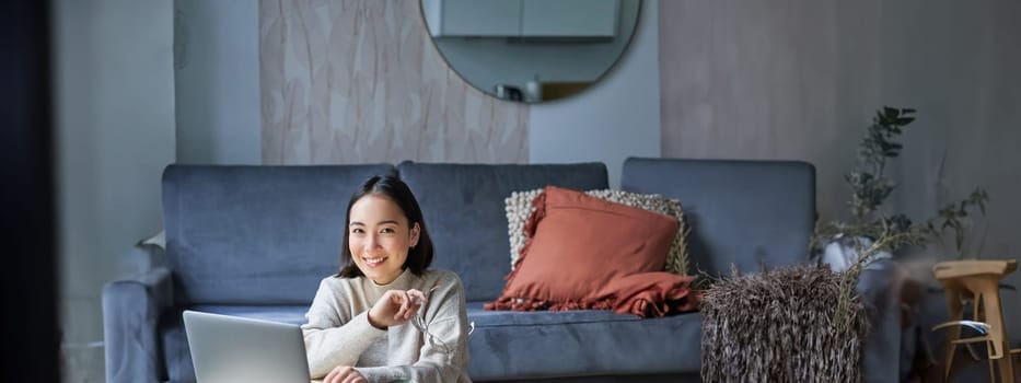 Vertical shot of asian girl sits on floor at home, working on laptop, studying at cozy place, using computer to freelance.