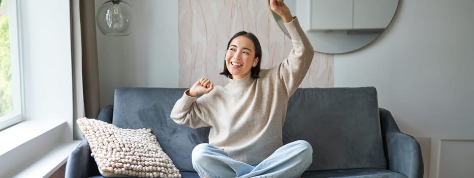 Happy and carefree girl singing and listening to music on smartphone app, using mobile phone as microphone, sitting on sofa.