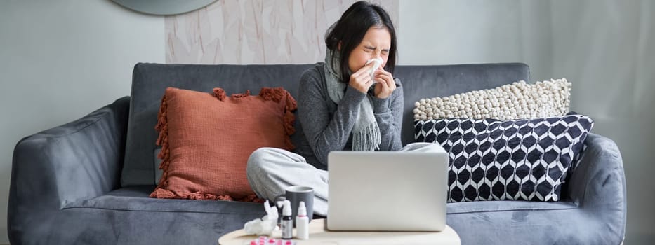 Young asian woman staying at home, feeling unwell, catching a cold, sick leave, sitting with laptop, watching movies on computer, sneezing.