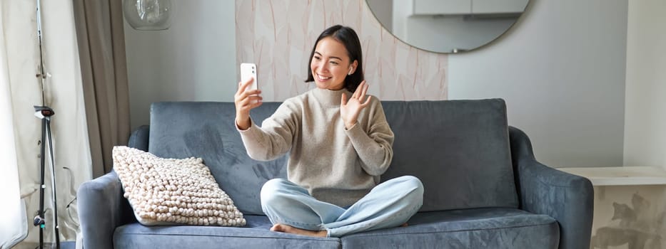 Portrait of asian woman sitting on sofa with smartphone, waving at mobile phone screen and waving at camera, video chat, talking to someone.