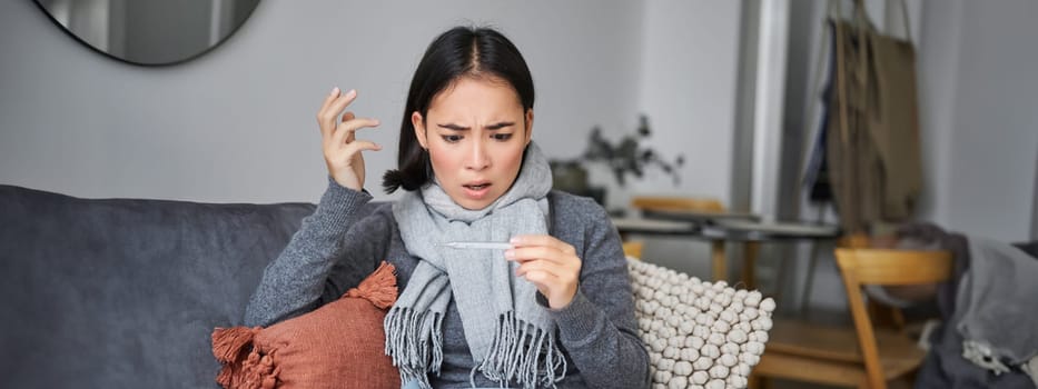 Shocked woman looks concerned at her thermometer, catching cold, has fever, sitting on sick leave at home.