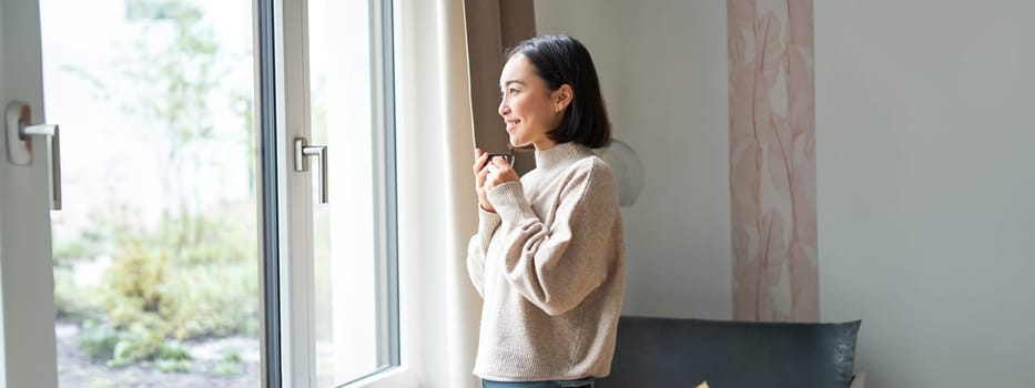 Beautiful korean woman staying at home, looking outside window, drinking coffee espresso and smiling, feeling comfort and warmth.