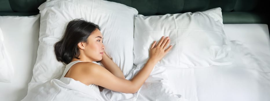 Portrait of romantic asian woman lying alone in her bed, touching empty pillow and thinking about her partner, breakup relatioship.