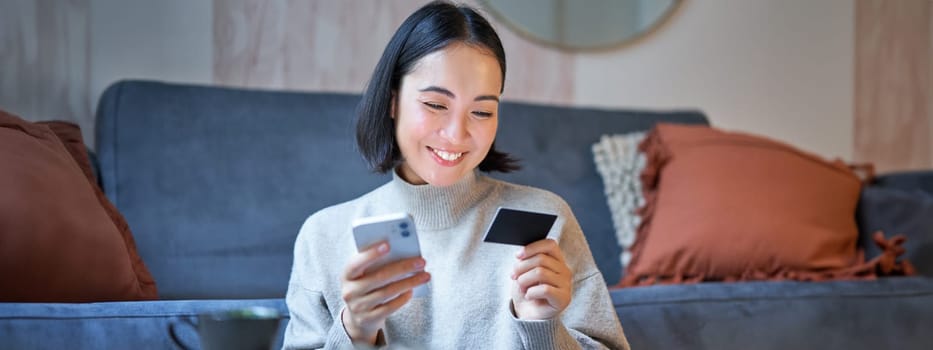 Portrait of smiling asian woman paying with credit card on her smartphone app, arrange direct debit or shopping online from mobile phone.