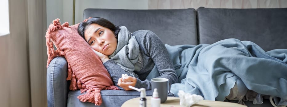 Sick sad korean woman lying on sofa, feeling unwell, catching cold, flu and temperature, looking upset, taking medication.