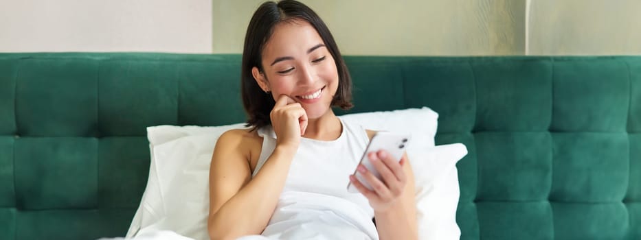 Beautiful korean woman reading mobile phone, messaging or watching video while relaxing in her bedroom, holding smartphone.