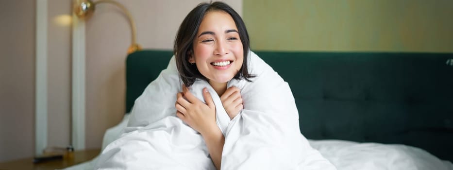 Cute and tender woman sitting in bed with warm duvet, smiling as looking out of the window, enjoying morning and comfort of blanket.