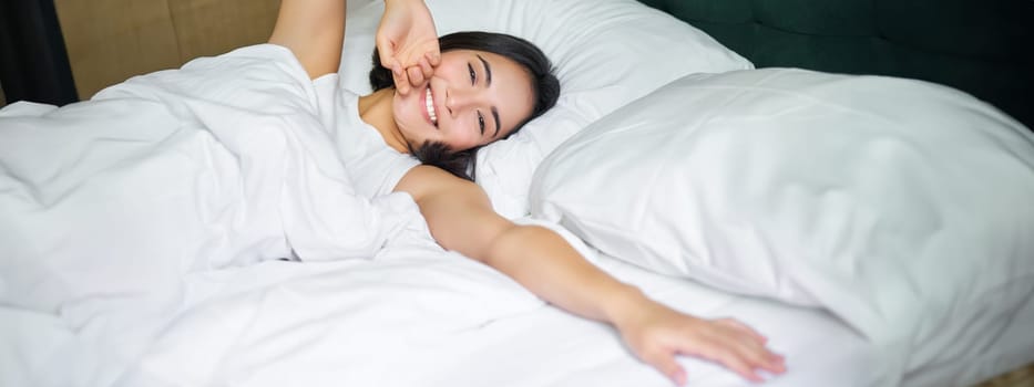 Romantic asian girl waking up in cozy bedroom, lying in bed with white sheets, stretching her hand towards empty pillow.