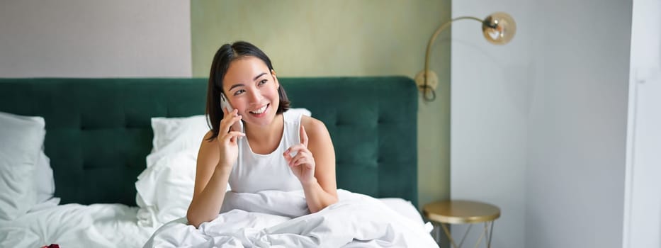 Beautiful young korean woman calling, having a phone call in bed, talking with friend while staying in bedroom.