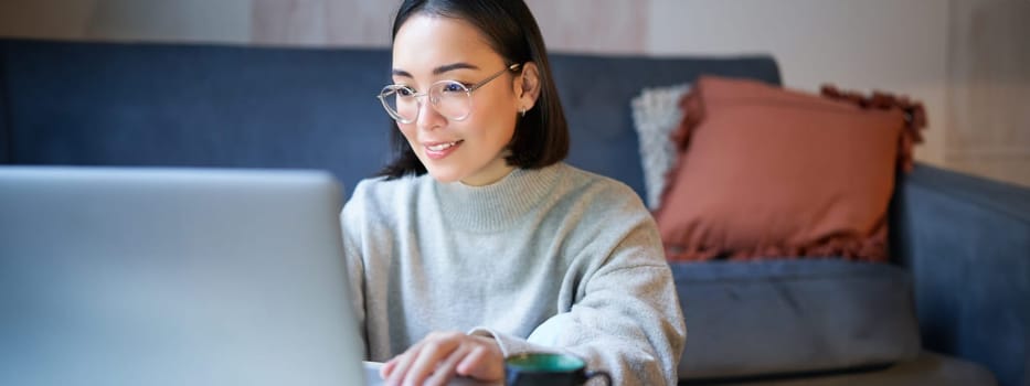 Portrait of smiling asian girl working from home, staying on remote, using laptop, studying on her computer.