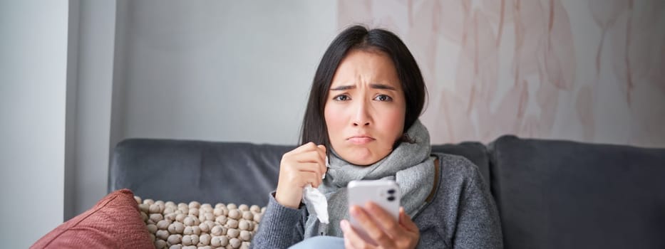 Asian girl with influenza, sits at home, cries and looks upset, holds smartphone, feels unwell and gloomy, wearing warm clothes.