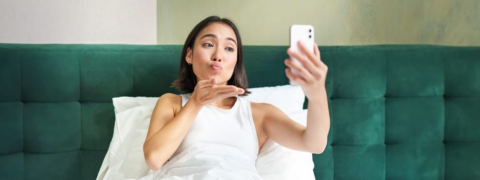 Beautiful asian girl lying in bed, making morning selfie, taking picture on smartphone in bedroom, smiling happily.