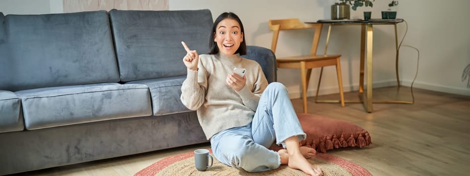 Smiling asian girl sits on floor in stylish living room, pointing finger at advertisement, showing promo banner, holding mobile phone in hand.