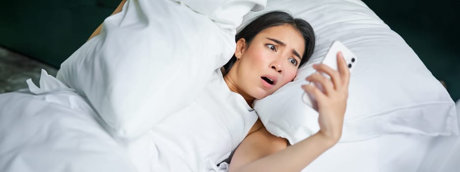 Portrait of asian woman waking up in bed, looking shocked at mobile phone, realise she overslept.