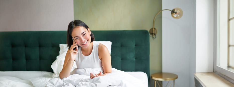 Smiling korean girl talks on mobile phone and lying in bed. Cute woman answers telephone call, holds smartphone, relaxing in her bed.