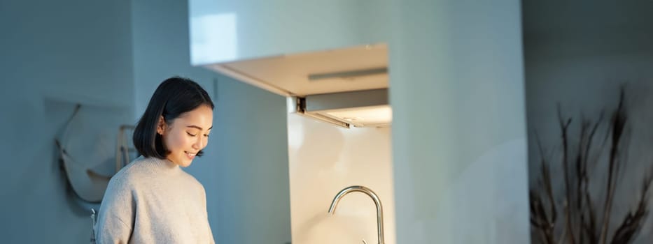 Vertical shot of young asian woman cooking dinner, making herself sandwitch, smiling while standing on the kitchen.