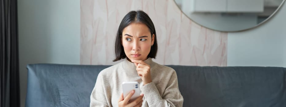 Portrait of girl sitting on sofa with smartphone, looking thoughtful and hesitant at mobile phone screen.