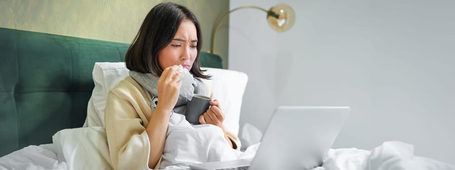 Portrait of asian girl sneezing and wiping runny nose, feeling sick, staying in bed with influenza, looking at laptop.