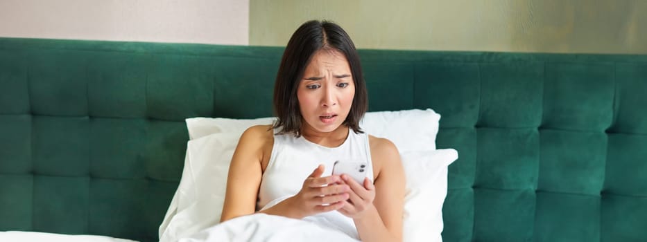 Bedroom picture of asian woman lying in bed, looking scared and shocked at smartphone screen, reading message with concerned face.