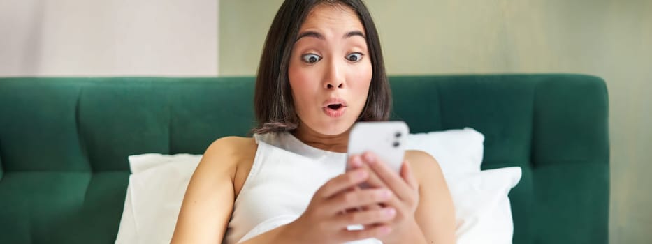 Cute asian girl holding smartphone, looking surprised at mobile phone screen, reading message with amazed face expression.