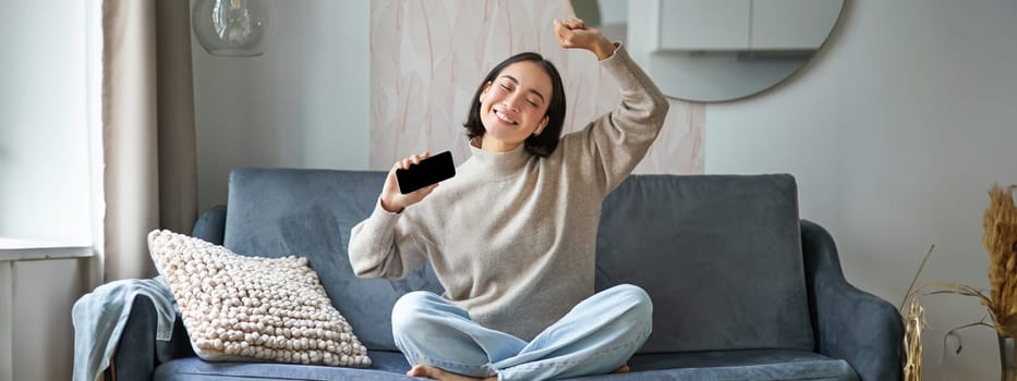 Happy and carefree girl singing and listening to music on smartphone app, using mobile phone as microphone, sitting on sofa.