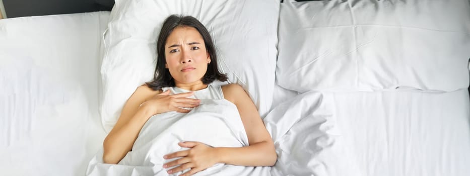 Portrait of sad young asian woman lying in bed, overthinking before going to sleep, stay late because of insomnia, frowning and looking frustrated.