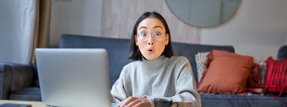 Portrait of asian woman in glasses, sitting with laptop and looking surprised, amazed by promotion on computer.