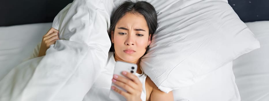 Close up portrait of asian girl lying in bed, looking at smartphone concerned, waking up late and staring at her alarm clock on mobile phone.