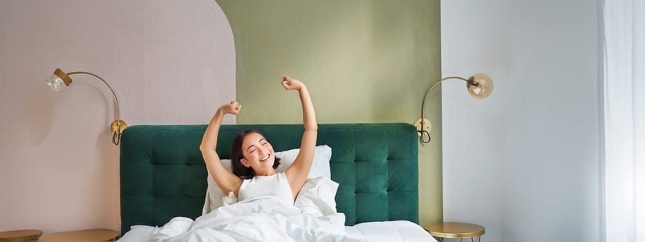 Portrait of smiling happy asian girl, wakes up feeling enthusiastic, stretches her hands up, enjoys good morning in her bedroom.