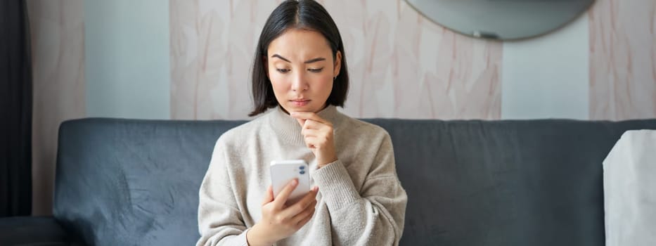 Portrait of girl sitting on sofa with smartphone, looking thoughtful and hesitant at mobile phone screen.