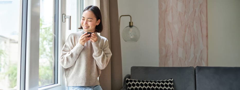 Portrait of young asian woman sitting near window and looking outside, drinking hot coffee from espresso cup and enjoying her cozy day off at home.