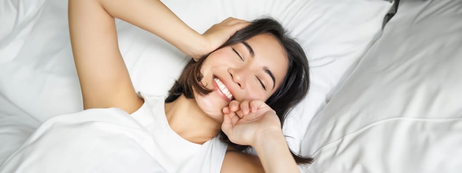 Close up of happy asian girl lying in bed on her pillow, smiling and rolling over as feeling energetic in morning, waking up early.