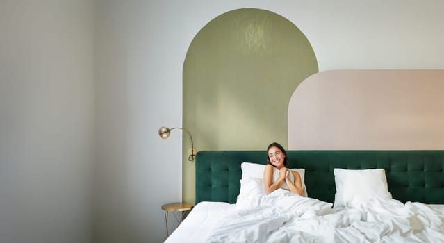 Vertical shot of beautiful asian girl lying in bedroom with sophisticated interrior, smiling and looking happy in morning, spending time in bed.