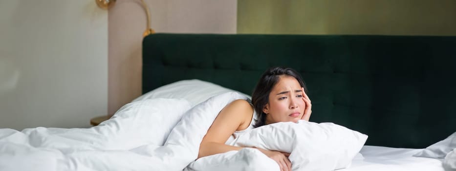 Sad and distressed young korean woman lying in her bed with pillow and warm duvet, feels uneasy, frowning and grimacing.