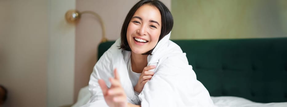 Happy beautiful asian woman covered with blanket, sits on bed in warm duvet, tries to reach camera with a hand, smiling playfully. Hotel bedroom, comfort and apartments concept.