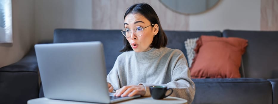 Portrait of asian woman in glasses, sitting with laptop and looking surprised, amazed by promotion on computer.