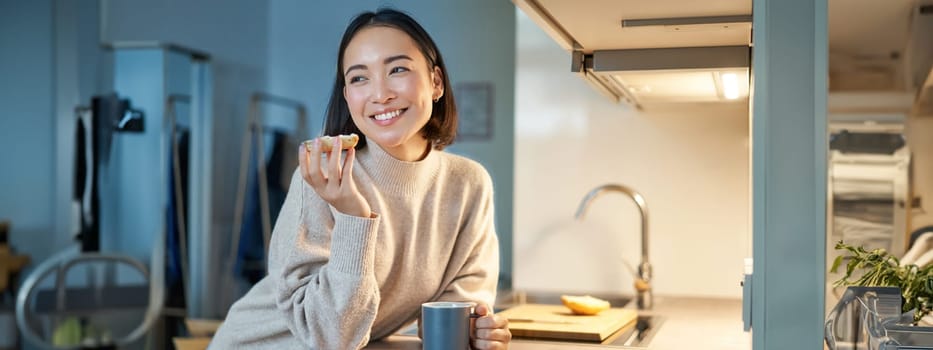 Beautiful young asian woman eating toast on her kitchen, drinking coffee and smiling.