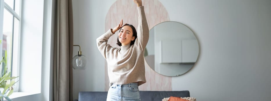 Women and wellbeing. Beautiful young asian woman dancing, feeling carefree and happy, raising her hands above head and smiling, enjoying her stay at home.