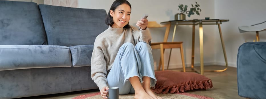 Portrait of beautiful asian girl sitting at her home and watching tv, holding remote, smiling and laughing, feeling comfort and warmth at her apartment.