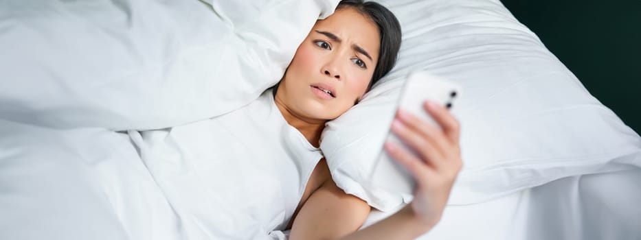 Portrait of asian woman waking up in bed, looking shocked at mobile phone, realise she overslept.