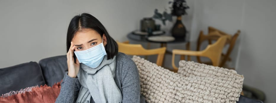 Covid-19 and health concept. Young asian woman in medical face mask, feels sick and unwell, catching flu, protecting others from influenza, sitting in living room covered with blanket.
